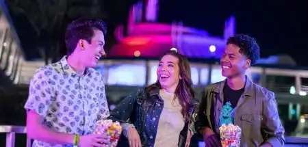 Three friends smiling and enjoying snacks together outdoors at night with colorful lights in the background