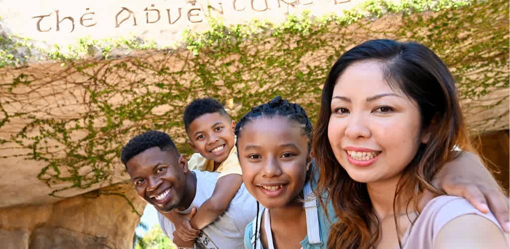 A diverse group of smiling children and a woman taking a selfie outdoors under an archway with green vines.