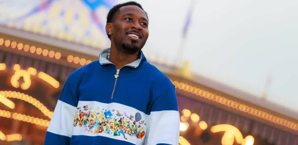 A smiling man standing outdoors at a carnival with bright lights and a ferris wheel in the background during the evening.