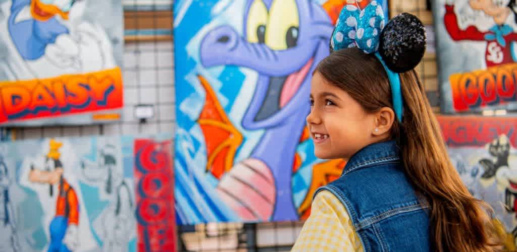 A young girl with long brown hair wearing Mickey Mouse ears smiling at colorful cartoon character art displayed outdoors