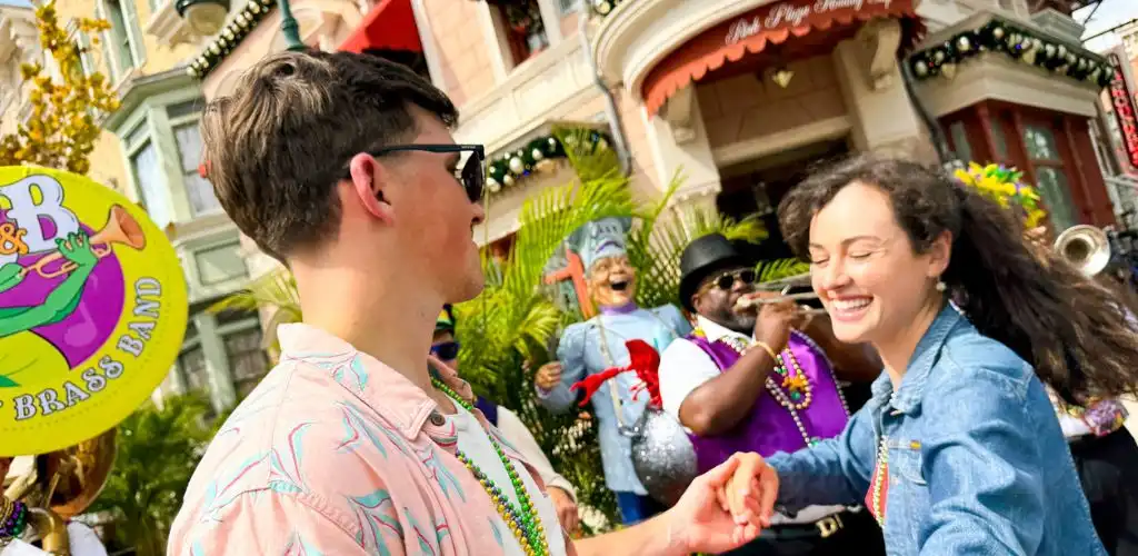 A smiling young woman and a young man hold hands while dancing outdoors during a lively parade with musicians and colorful decorations.