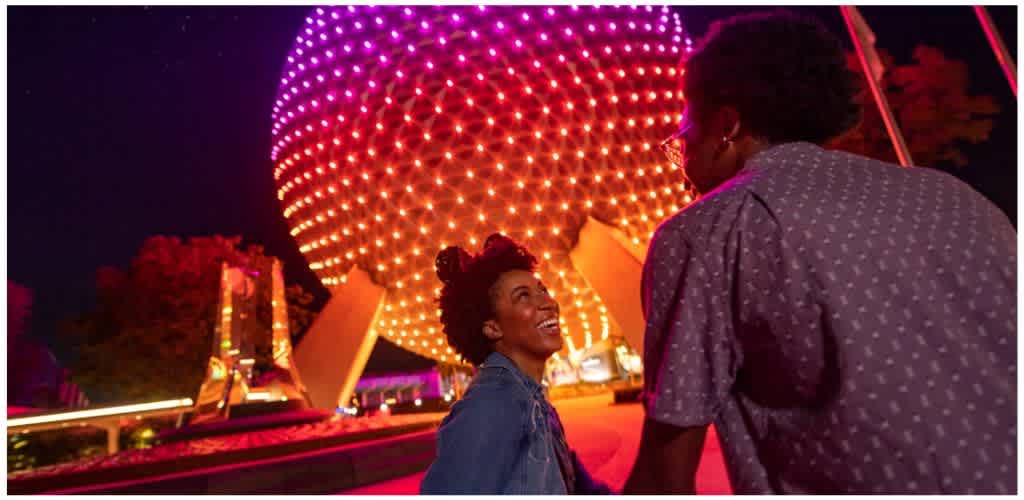 Two people smiling and talking at night in front of a brightly lit spherical-shaped structure with colorful lights.