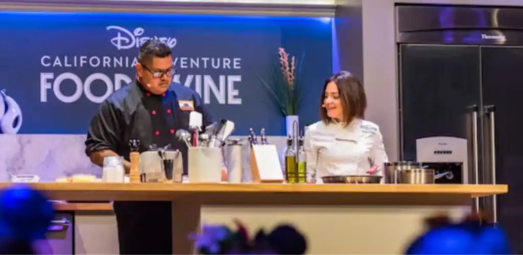 A man and woman in chef uniforms are preparing food in a kitchen studio with a Disney California Adventure Food & Wine sign in the background.