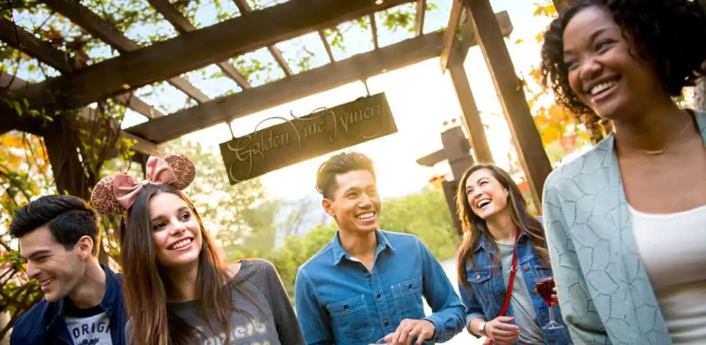 Group of diverse young adults smiling outdoors at a gathering under a wooden pergola with a sign that says Golden Vibe Vineyard