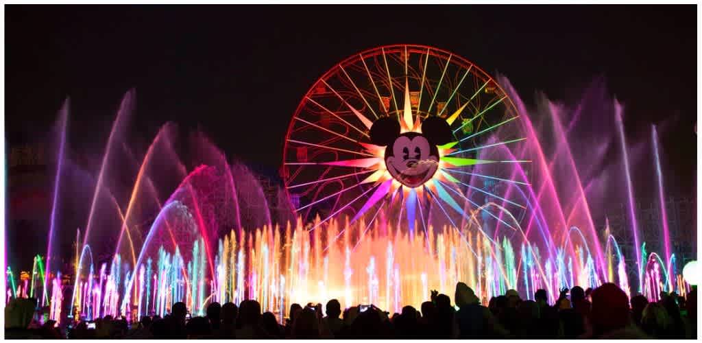 Colorful nighttime water fountain display at a theme park with a large Mickey Mouse face and Ferris wheel in the background