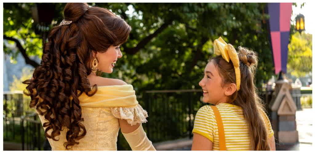 A woman dressed as Belle from Disney's Beauty and the Beast smiling and talking with a young girl in a yellow striped shirt outdoors.