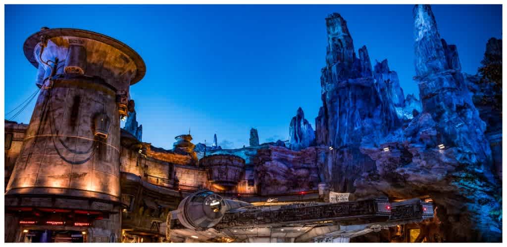 A futuristic manufacturing building with rocky cliffs and blue-lit mountains in the background at night.