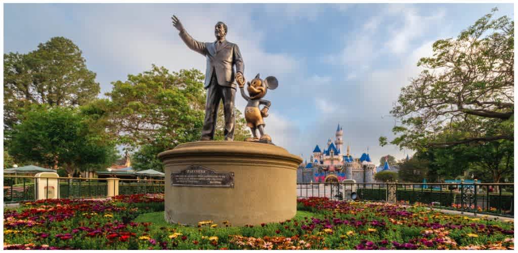 Statue of Walt Disney and Mickey Mouse standing on a circular pedestal with a castle in the background and colorful flowers in the foreground