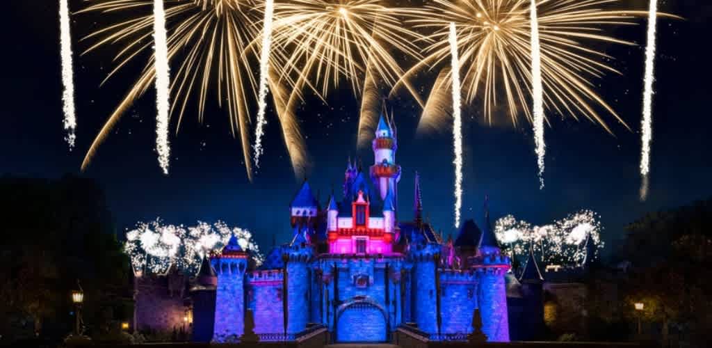 A brightly lit Disney castle at night with colorful fireworks illuminating the sky above it.