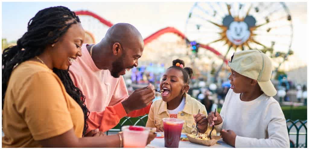 A family enjoying a meal outdoors at an amusement park with a Ferris wheel and a smiling sun decoration in the background.