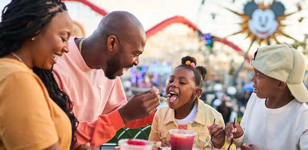 A family enjoying a meal together outdoors at an amusement park with a Ferris wheel and Mickey Mouse in the background.