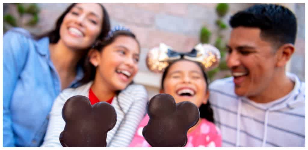 Four people smiling and holding Mickey Mouse-shaped cookies in front of a blurred outdoor background.