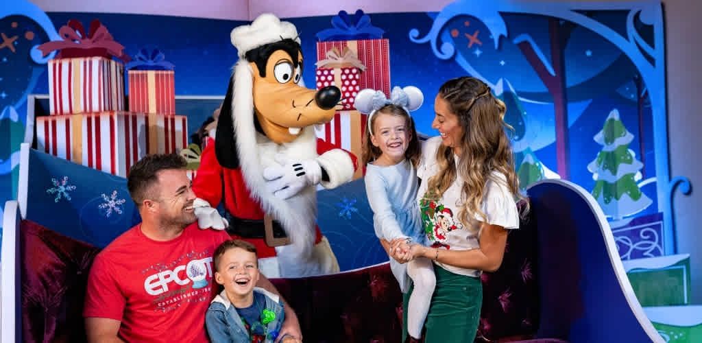 A smiling family with a child on Santa's sleigh, joined by a person in a Mickey Mouse costume, and a festive holiday background with presents and decorated trees.