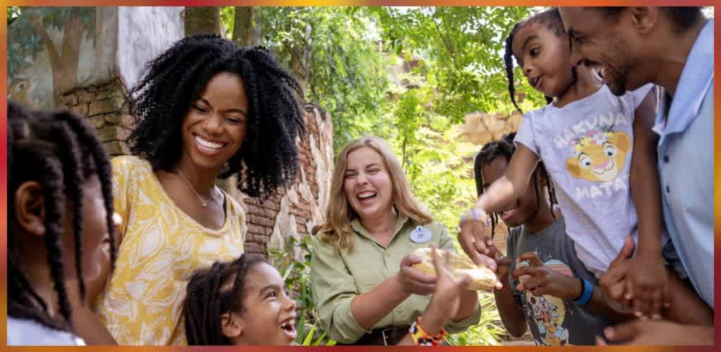 A diverse group of children and adults smiling and sharing food outdoors surrounded by trees and greenery.