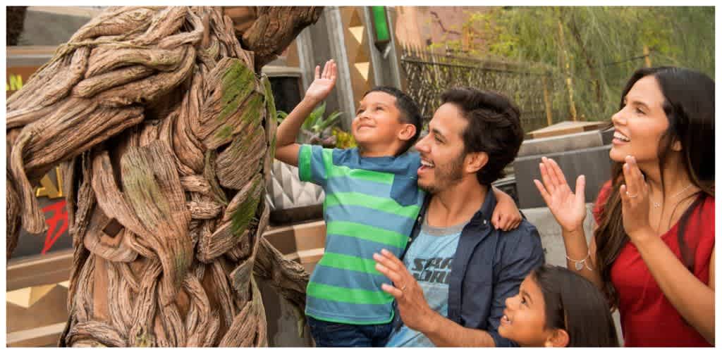 A family smiling and waving at a person in a tree costume outdoors with trees and a wooden fence in the background.