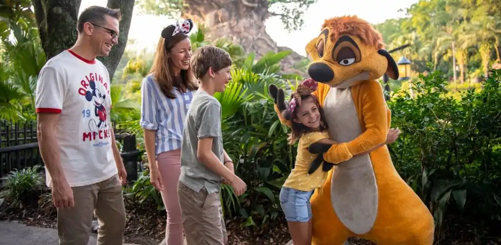 Two children are smiling with two Disney character mascots in a lush outdoor setting with green plants and trees.