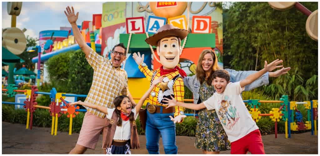 Happy family with two children posing with Woody from Toy Story at an amusement park with colorful rides and attractions.