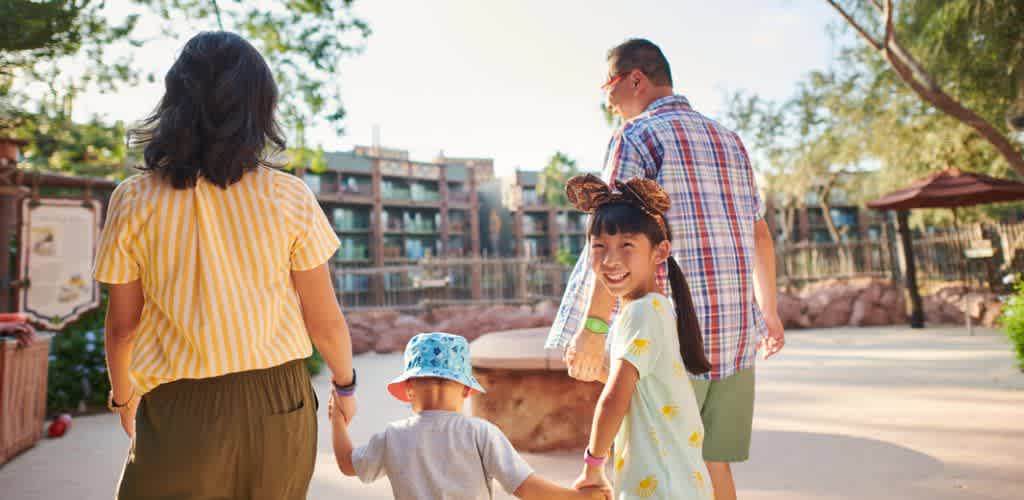 A family enjoying a sunny day at the park with children holding hands and smiling, surrounded by trees and playground structures.