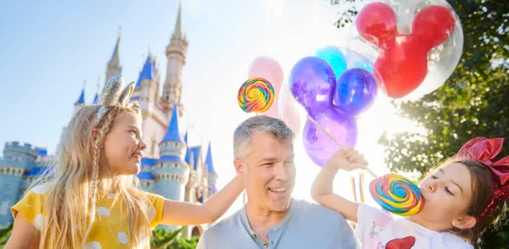 A man and two young girls celebrate at a castle park with colorful balloons and large lollipops on a sunny day.