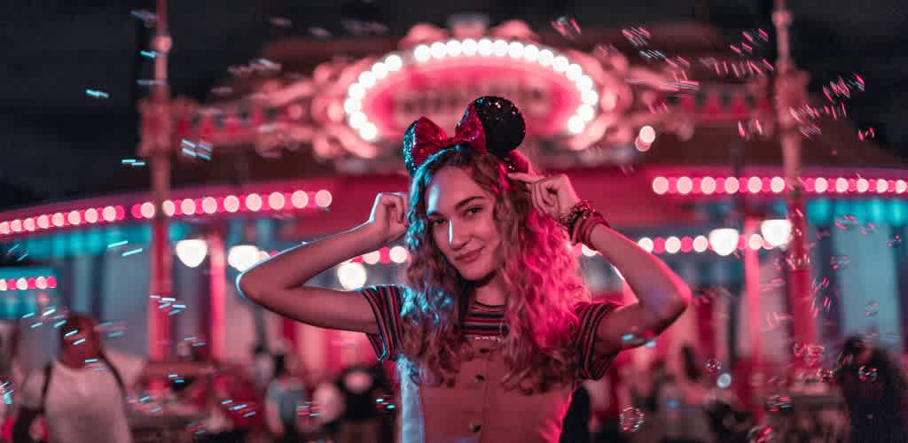 Teen girl wearing Minnie Mouse ears at a colorful amusement park nighttime event smiling at the camera