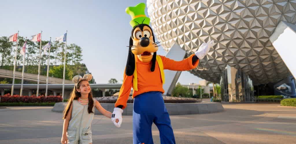 A young girl holding hands with a person in a Pluto mascot costume outside a theme park with a spaceship earth-like building in the background.