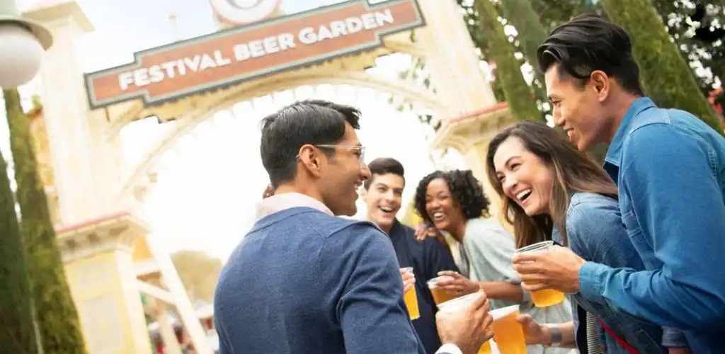 Group of diverse people smiling and enjoying drinks at a festival beer garden outdoors with trees in the background