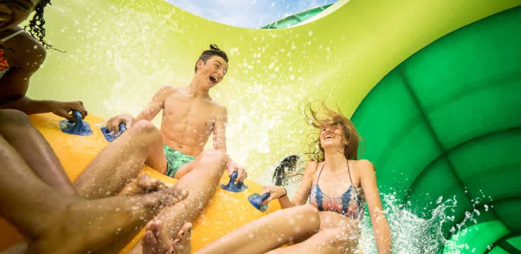Children having fun on an outdoor water slide with bright yellow and green inflatable sides and splashing water.
