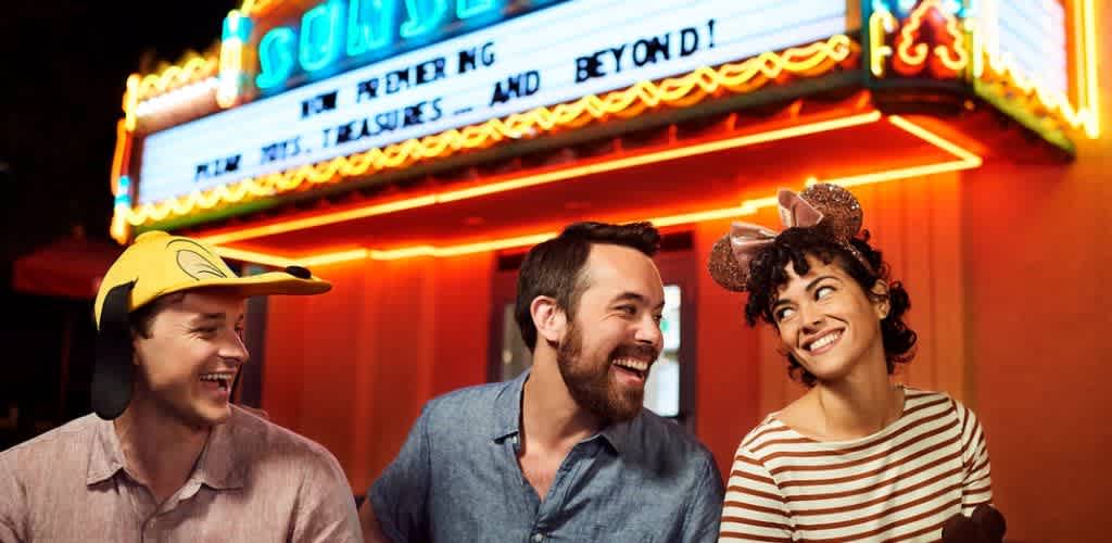 Three people smiling and enjoying themselves at an amusement park with colorful lights and a marquee sign in the background.