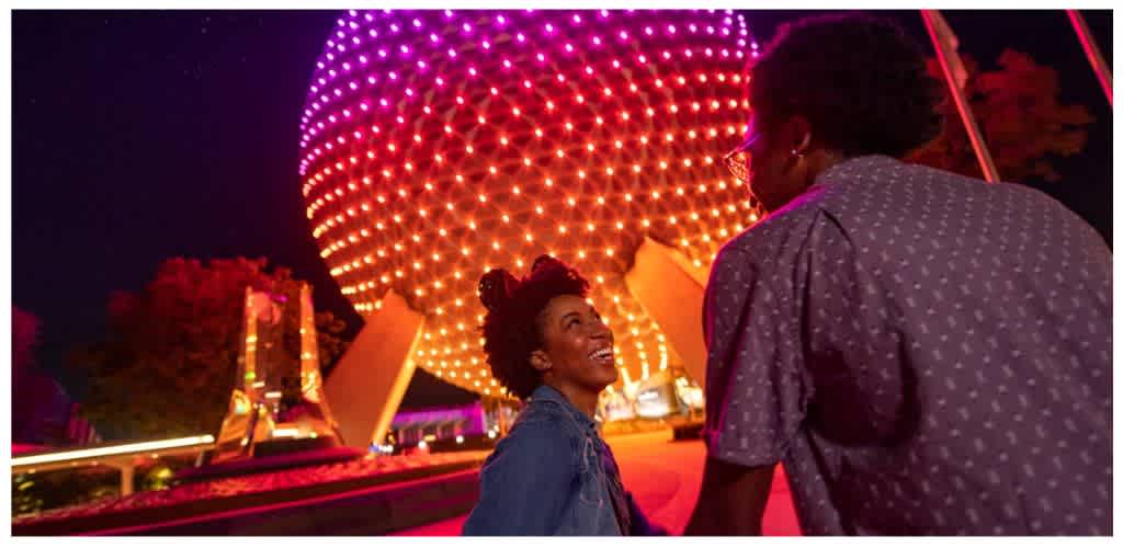 Two people talking near a brightly lit spherical attraction at night in an amusement park