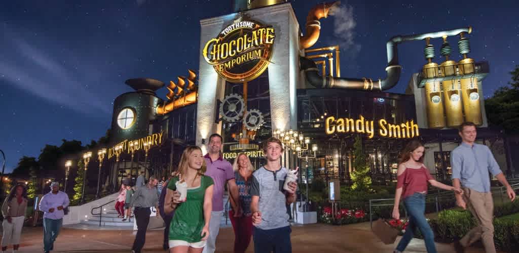 People walking outside a themed chocolate factory building at night with illuminated signs and steampunk-inspired architecture