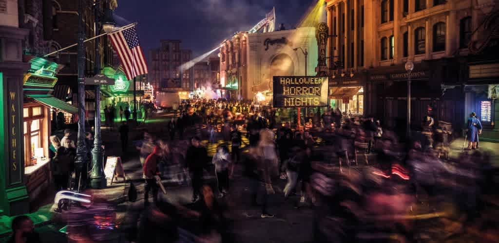 Night scene of a busy street with blurred crowds, colorful lights, and a sign for Halloween Horror Nights.