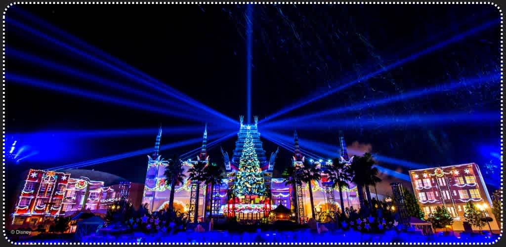 Nighttime scene of a decorated building with colorful lights, a large Christmas tree in front, palm trees, and bright blue laser beams lighting the sky.