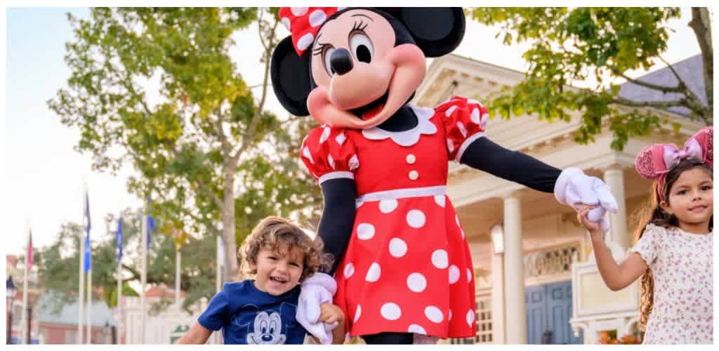 Minnie Mouse character dressed in red and white polka dot dress holding hands with children outdoors at a theme park