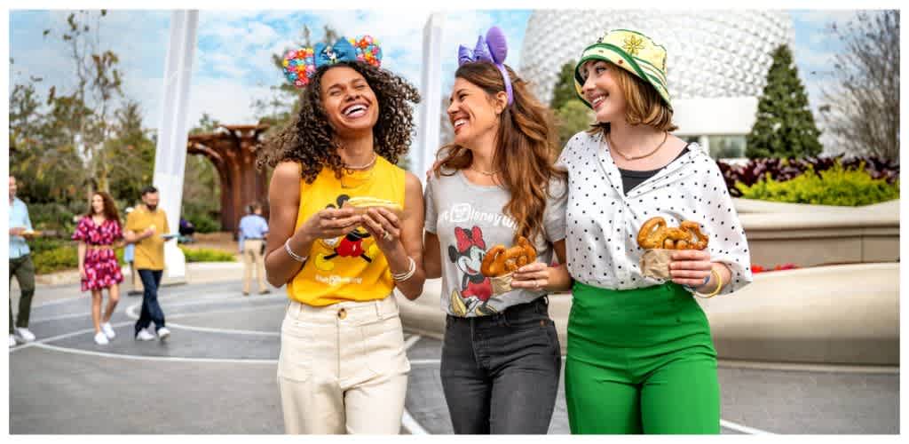 Three women smiling and holding pretzels in an outdoor park with a modern building and other people in the background