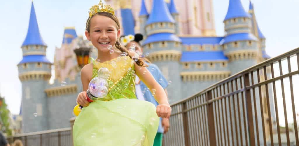A young girl dressed as a princess smiles happily at a castle in the background while holding a bubble wand.