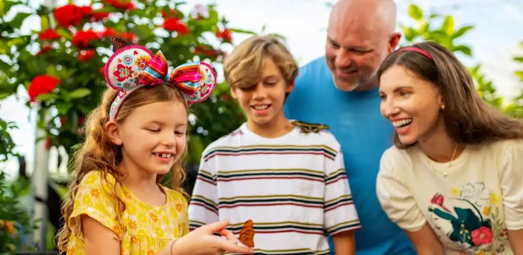 A family of four happily smiling and looking at a small butterfly outdoors in a lush garden setting.