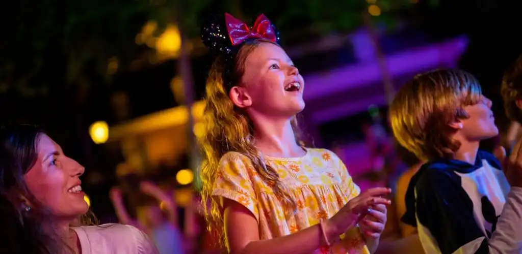 A smiling girl with a bow headband enjoying an outdoor event at night with colorful lighting and other children around her.