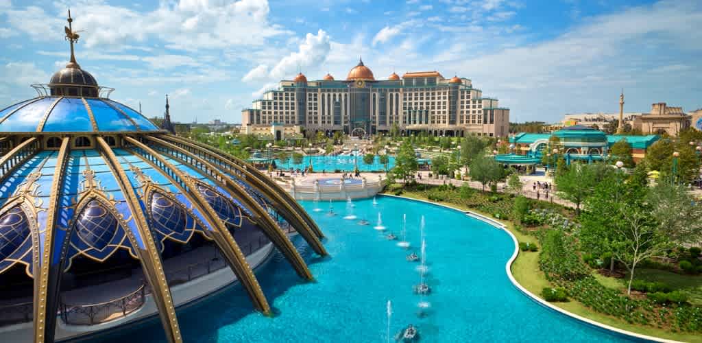Aerial view of a luxurious resort with a domed structure, fountains, lush green landscaping, and a large hotel building in the background under a blue sky
