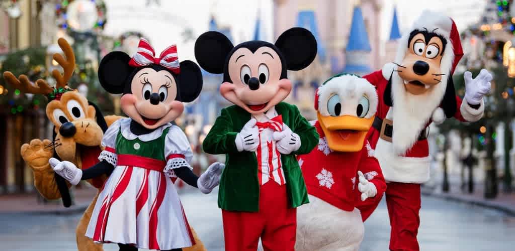 Disney characters dressed in holiday costumes parade outdoors on a festive decorated street with blurred buildings in the background.