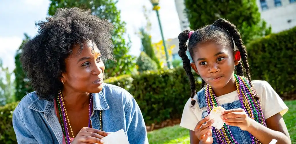A woman and girl sitting outdoors smiling and eating ice cream during a festive event with colorful beads and greenery in the background.