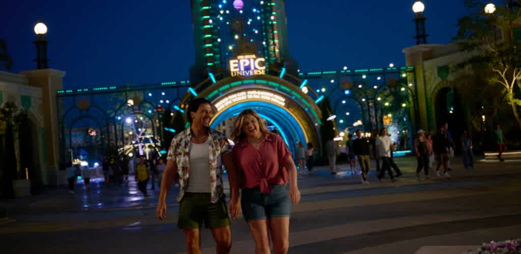 Two women walk happily under an illuminated amusement park entrance at night with colorful lights and crowds around.