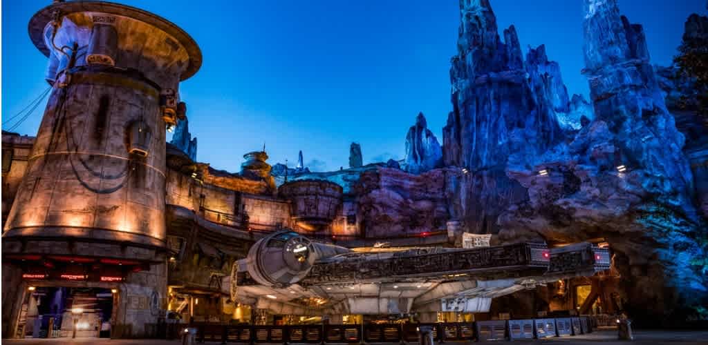 A futuristic spaceship is parked outside a rocky, space-themed building with tall, jagged blue mountains in the background during evening or night.