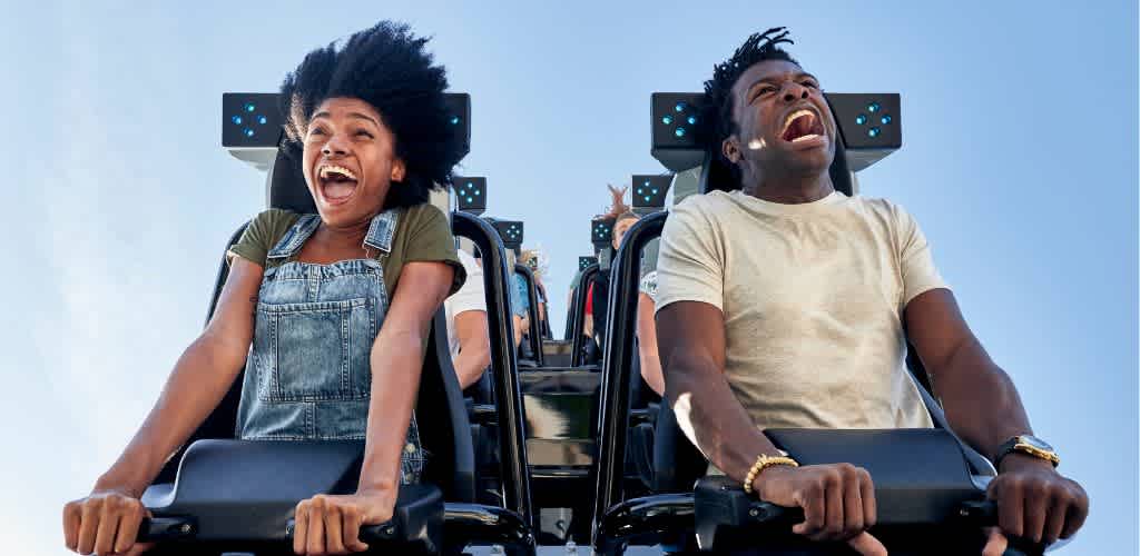 Two people are enjoying a roller coaster ride, smiling and laughing with their hands on the safety bars against a clear blue sky.