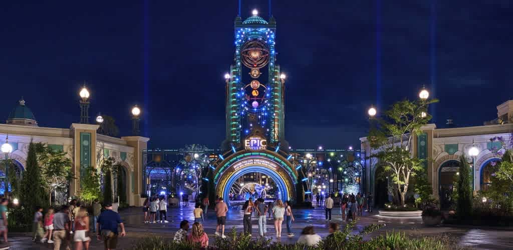 Nighttime view of an amusement park entrance with colorful lights, a tall tower, and visitors walking inside.