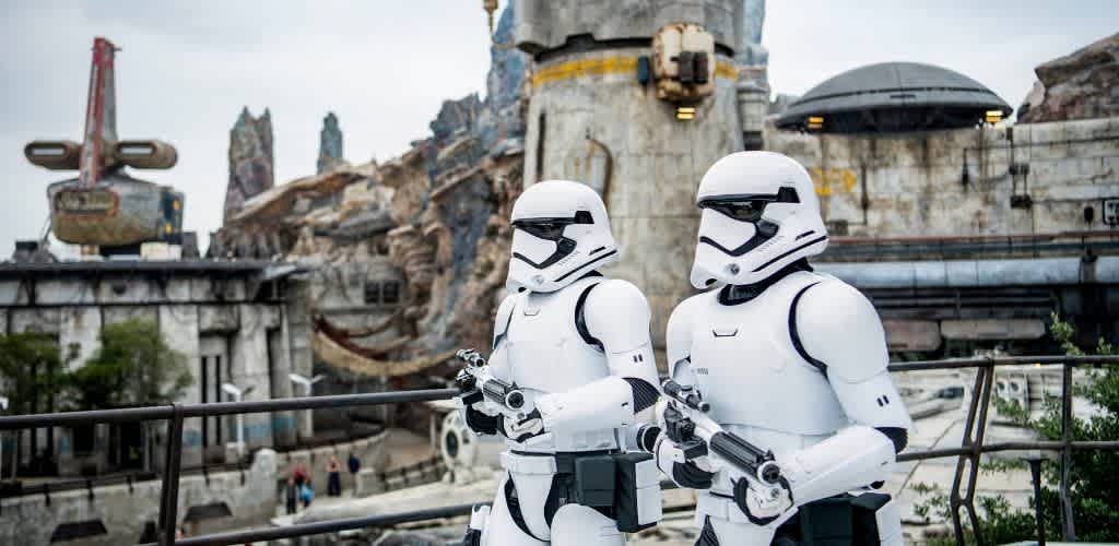 Two stormtroopers stand outdoors in front of a science fiction landscape with rocks, structures, and a spaceship in the background.