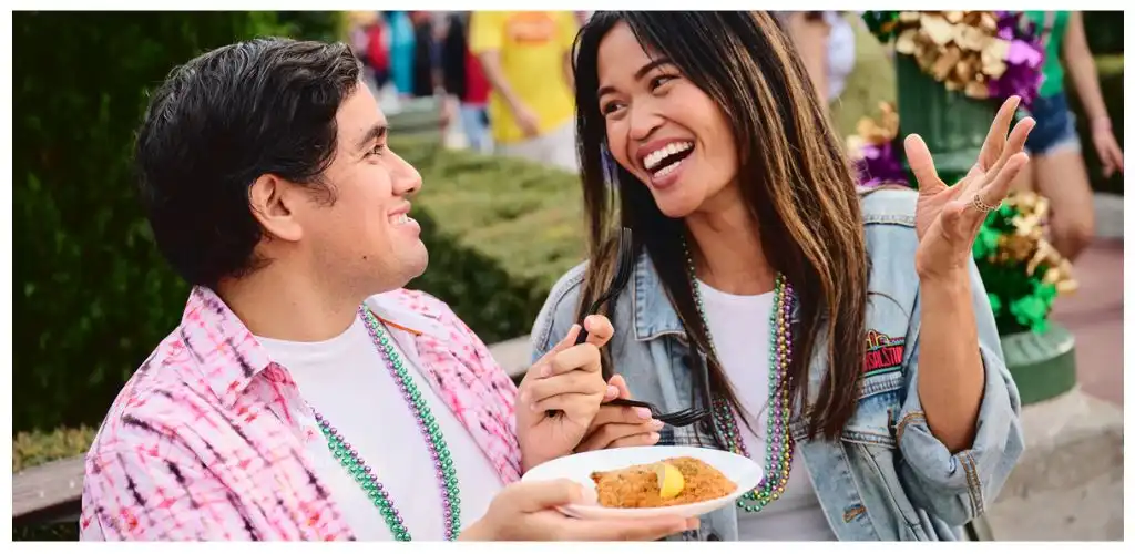 Two people smiling and talking outdoors during a celebration, holding a plate of food and wearing colorful bead necklaces.