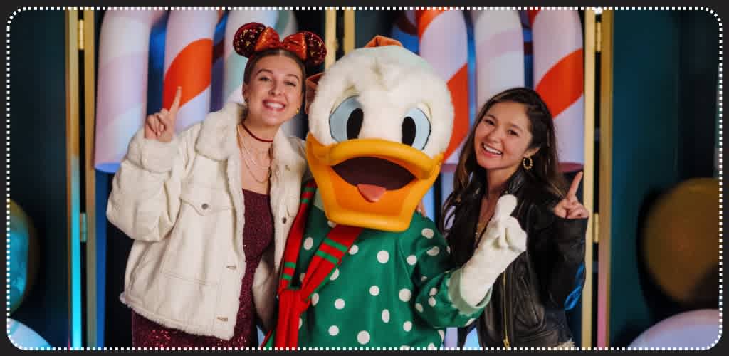 Two women and a Donald Duck mascot posing together at a festive event with colorful holiday decorations in the background