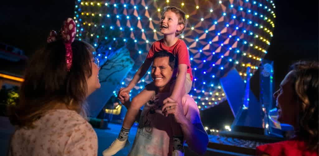 A smiling man carries a child on his shoulders while two women look on happily at an illuminated amusement park night scene with colorful lights