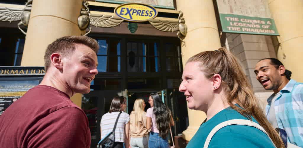Group of people smiling and conversing outside the entrance of a theme park or attraction with a sign about the pyramids visible in the background