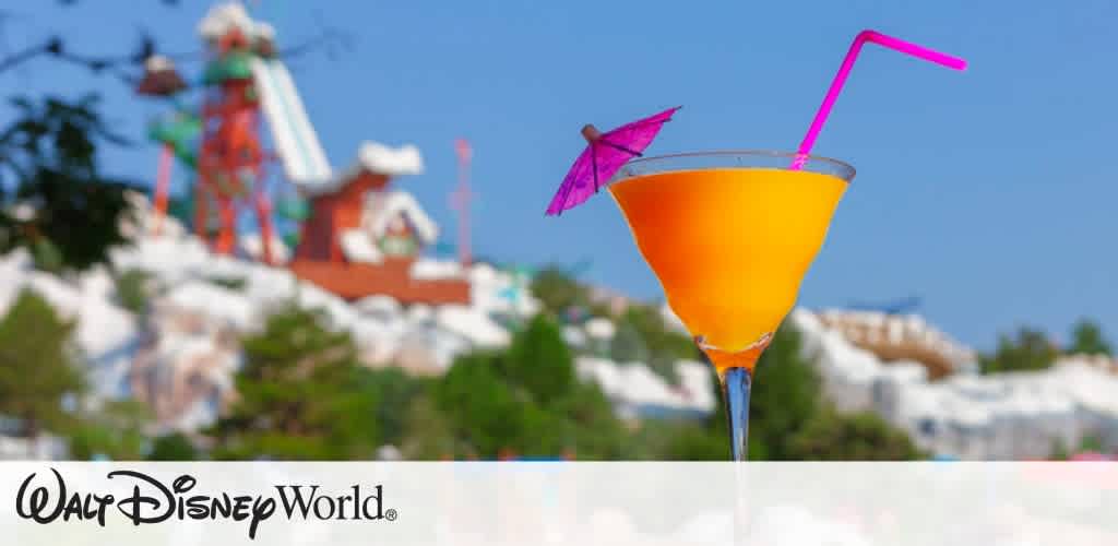 A vibrant, tropical cocktail is prominently featured in the foreground, showcasing a bright orange drink adorned with a small pink umbrella and a pink straw. In the blurred background, iconic water slides and green trees suggest a lively atmosphere at a Walt Disney World water park under a clear blue sky. The image conveys a fun, relaxing vacation vibe.